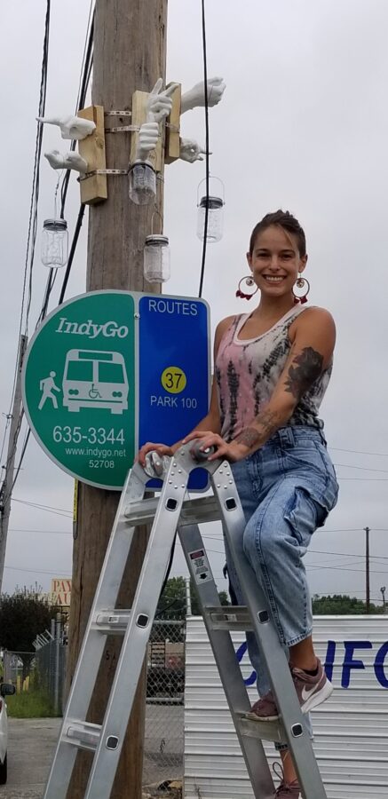 woman on ladder poses with clay hand figures above bus sign